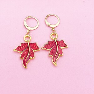 May include: A pair of gold earrings with red leaf-shaped charms. The earrings have a hoop design and are perfect for adding a touch of nature to any outfit.