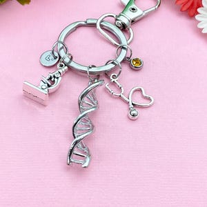 May include: Silver-toned keychain featuring medical-themed charms. Charms include a microscope, DNA helix, and a stethoscope shaped like a heart. The keychain also has a small heart charm and a yellow gemstone. The background is a light pink color.