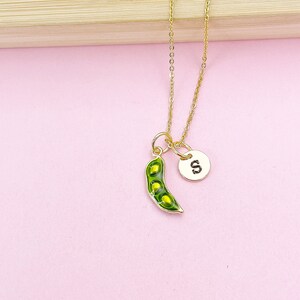 Peut inclure: Un collier dor&eacute; avec un pendentif en forme de gousse de pois verte contenant trois pois jaunes, et un petit charme rond avec la lettre "S". Fond rose.