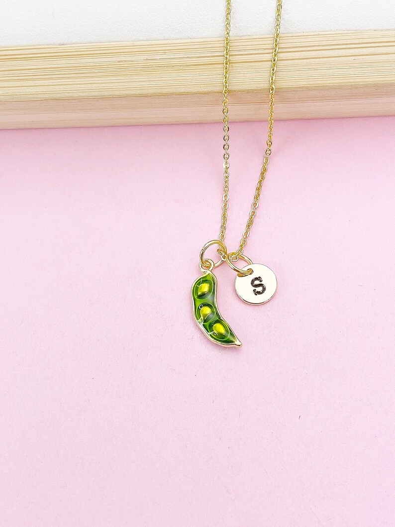 Peut inclure: Un collier dor&eacute; avec un pendentif en forme de gousse de pois verte contenant trois pois jaunes, et un petit charme rond avec la lettre "S". Fond rose.