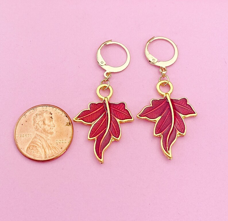 May include: A pair of gold earrings with red enamel leaf designs. The leaves are detailed with gold veins and have a slightly textured surface.