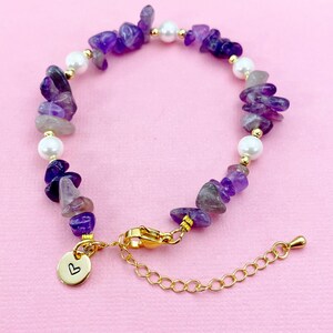 May include: A beaded bracelet featuring amethyst chips, white pearls, and gold-colored accents. The bracelet has a gold-colored clasp and a small charm with a heart symbol. The jewelry is displayed on a pink background.