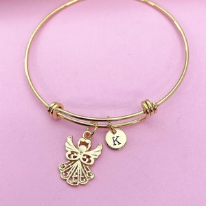 K&ouml;nnte beinhalten: Goldenes Armreif-Armband mit einem Engelsanh&auml;nger und einem kleinen runden Anh&auml;nger mit dem eingravierten Buchstaben "K".