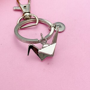 K&ouml;nnte beinhalten: Silberner Origami-Kranich-Schl&uuml;sselanh&auml;nger mit einem silbernen geteilten Ring und einem silbernen Karabinerhaken. Der Schl&uuml;sselanh&auml;nger hat auch eine kleine silberne Scheibe mit dem eingravierten Buchstaben "M".