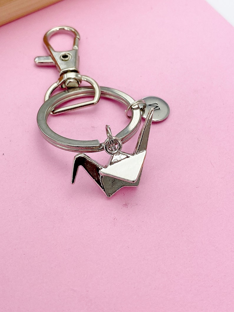 K&ouml;nnte beinhalten: Silberner Origami-Kranich-Schl&uuml;sselanh&auml;nger mit einem silbernen geteilten Ring und einem silbernen Karabinerhaken. Der Schl&uuml;sselanh&auml;nger hat auch eine kleine silberne Scheibe mit dem eingravierten Buchstaben "M".