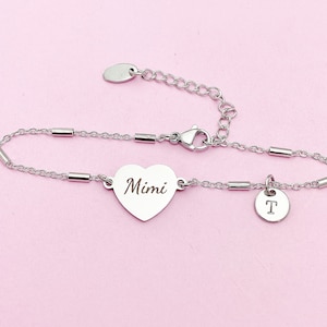 May include: A silver-colored bracelet with a heart-shaped charm engraved with the name "Mimi". The bracelet also features a small round charm with the letter "T". The bracelet is on a pink background.