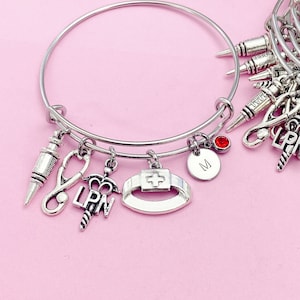 May include: A silver charm bracelet with a red stone charm and charms shaped like a stethoscope, a syringe, a nurse's cap, and a medical symbol. The bracelet also has a charm with the letters "LPN" on it.