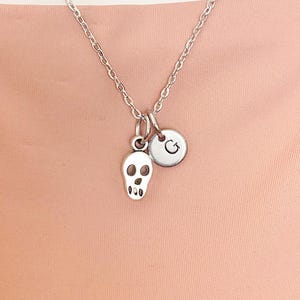 May include: A silver-toned necklace featuring a delicate chain with two charms attached: a small skull pendant and a circular disc charm engraved with the uppercase letter G. The metal components have a polished finish and are linked together.