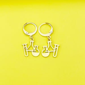 May include: A pair of gold-toned earrings with a dangle design featuring a beaker and test tube. The earrings are made of metal and have a hoop style.