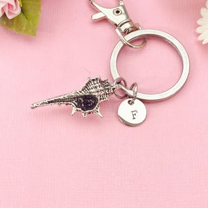 May include: Silver-toned keychain featuring a detailed seashell charm and a small round tag with the letter "F". The keychain has a lobster clasp and a large ring. The background is a soft pink.