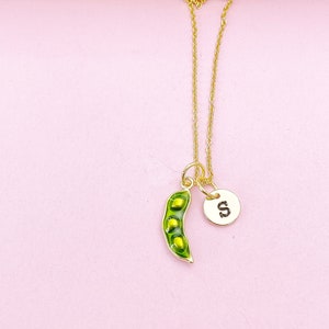 Peut inclure: Un collier dor&eacute; avec deux breloques. L'une est une gousse de pois verte avec des pois jaunes, l'autre un disque dor&eacute; avec la lettre "S". Le collier est pos&eacute; sur un fond rose. La breloque en forme de gousse mesure environ 2,5 cm de long.