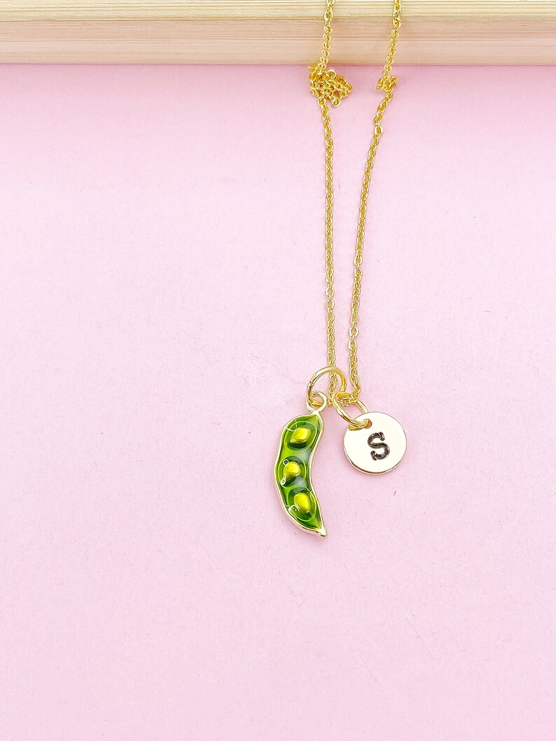 Peut inclure: Un collier dor&eacute; avec deux breloques. L'une est une gousse de pois verte avec des pois jaunes, l'autre un disque dor&eacute; avec la lettre "S". Le collier est pos&eacute; sur un fond rose. La breloque en forme de gousse mesure environ 2,5 cm de long.
