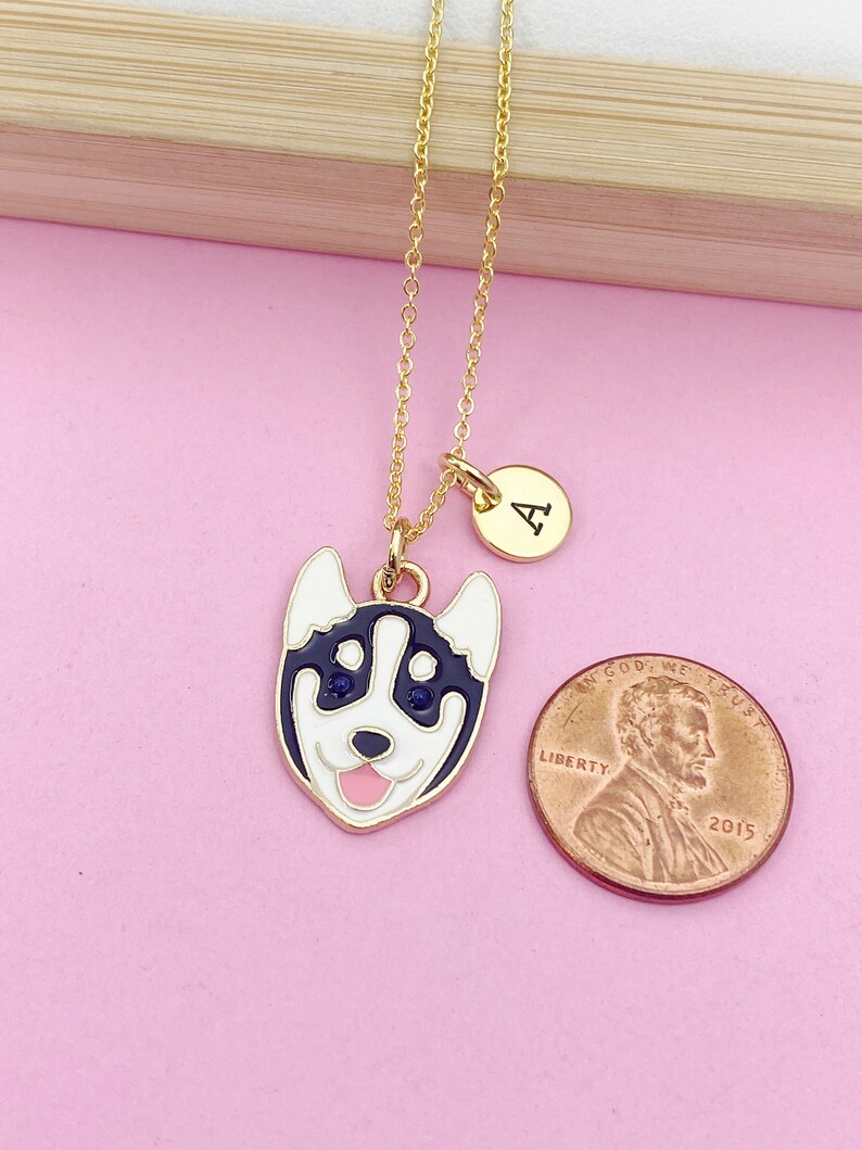 May include: Gold chain necklace with a charm of a cartoon husky dog with blue eyes and a pink tongue. The charm is attached to a gold circle with the letter "A" on it.