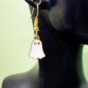 May include: A gold-toned metal earring featuring a small, white enamel ghost charm with two circular cutouts for eyes. The ghost charm hangs from a gold-toned fishhook ear wire, which includes a small gold bead and a coiled wire detail.
