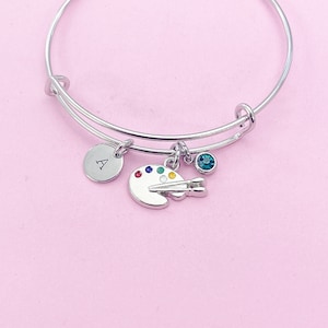 May include: Silver bangle bracelet with a charm of a paint palette and brush, a small blue gemstone, and a round charm with the letter A.