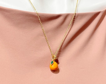 May include: A gold-colored chain necklace with a pendant shaped like a small orange fruit. The orange pendant has a green leaf and a small brown charm. The necklace is displayed against a pink background.