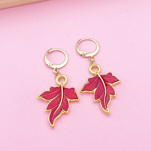May include: A pair of gold earrings with red leaf-shaped charms. The leaves have a detailed design and a glossy finish.