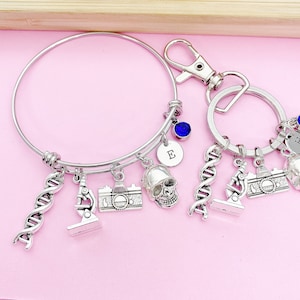 May include: A silver charm bracelet and keychain set with a variety of charms, including a skull, a microscope, a camera, and a DNA strand. The bracelet and keychain are both adorned with blue crystals and personalized with the letters "E" and "H".