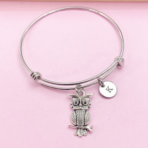 May include: A silver-colored adjustable bangle bracelet with an owl charm and a small round charm with the letter "K". The owl charm is detailed with textured feathers and perched on a branch. The background is a light pink.