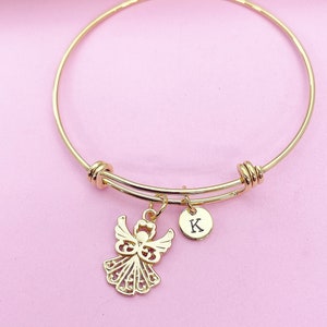 K&ouml;nnte beinhalten: Goldenes Armreif-Armband mit einem Engelsanh&auml;nger und einem kleinen Anh&auml;nger mit dem Buchstaben K. Das Armband liegt auf einem rosa Hintergrund.