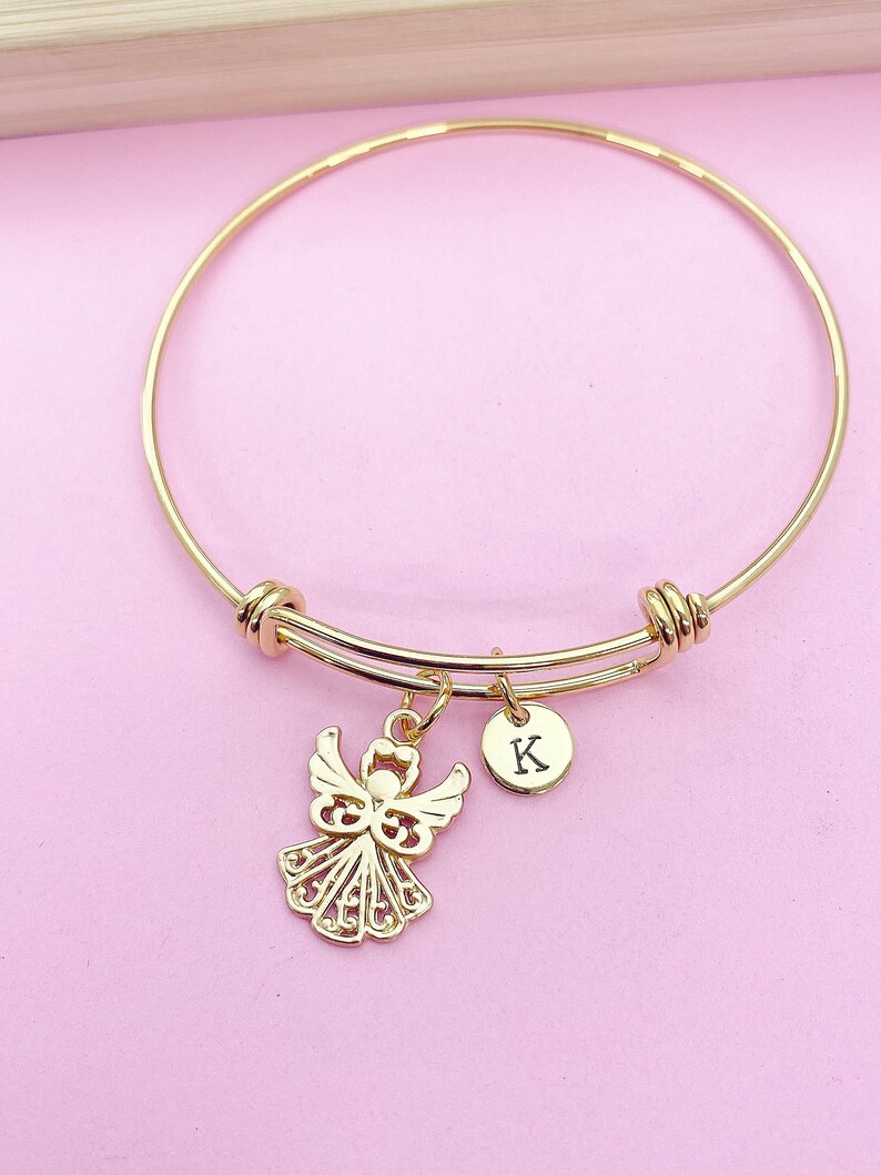 K&ouml;nnte beinhalten: Goldenes Armreif-Armband mit einem Engelsanh&auml;nger und einem kleinen Anh&auml;nger mit dem Buchstaben K. Das Armband liegt auf einem rosa Hintergrund.
