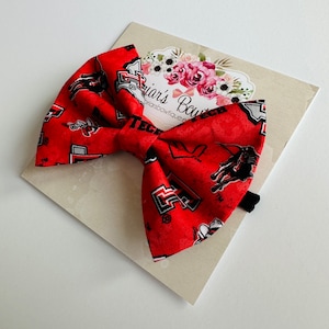 May include: Red and black bow with a Texas Tech University logo pattern. The bow is on a white background with a floral design.