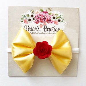 Puede incluir: Una diadema con lazo de satén amarillo con una rosa roja en el centro. El lazo está sujeto a una banda elástica blanca. El producto se muestra en una tarjeta con el texto "Briar's Boutique" y la URL de la tienda Etsy.