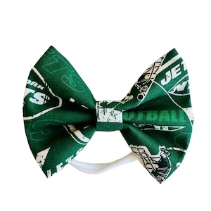 May include: Green and white bow headband with a New York Jets theme. The headband features a variety of logos and graphics, including the team's logo, the words "New York Jets", and the word "Football".