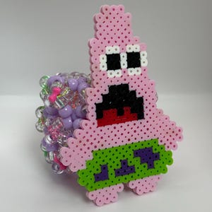 May include: A pink Patrick Star bead creation with black eyes and mouth, and a green and purple design on the bottom. The bracelet is attached to a beaded cuff with purple, pink, and clear beads.