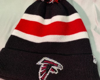 May include: Black knitted beanie with a white and red striped pattern and a black pom pom. The beanie features a rhinestone Atlanta Falcons logo on the front.