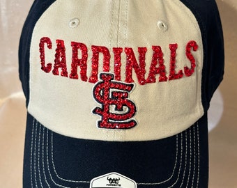May include: A white and black baseball cap with a red rhinestone Cardinals logo and the word "Cardinals" in red rhinestones. The cap is adjustable and has a "Fan Favorite" tag.