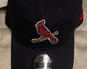 May include: A black baseball cap with a red cardinal bird embroidered on the front. The bird is covered in rhinestones and has a gold beak and feet. The cap has a black adjustable strap with a New Era logo and the text "9TWENTY ADJUSTABLE".