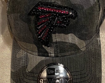 May include: Camouflage baseball cap with a black and red rhinestone Atlanta Falcons logo and a silver 9TWENTY adjustable label.