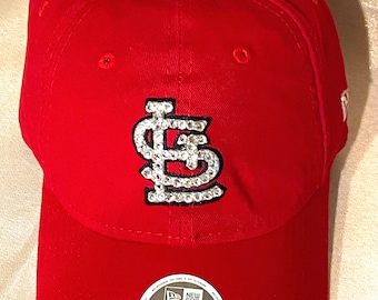 May include: A red baseball cap with a curved brim. The cap features a rhinestone-studded logo and a white and black tag that reads "9TWENTY WOMEN ADJUSTABLE".