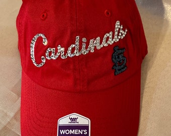 May include: Red baseball cap with a white rhinestone "Cardinals" logo and a black team logo. The cap has an adjustable strap with a "Fan Favourite Women's Adjustable" label.