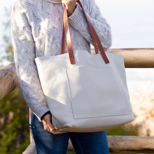 Watch the Tote Glo Bag. This tote bag is extremely light, the leather is ultra soft, buttery, very durable and resistant.
This bag is white, have tan leather handles and outside pocket. The bag appears grabbed by a model in front of wooden logs.