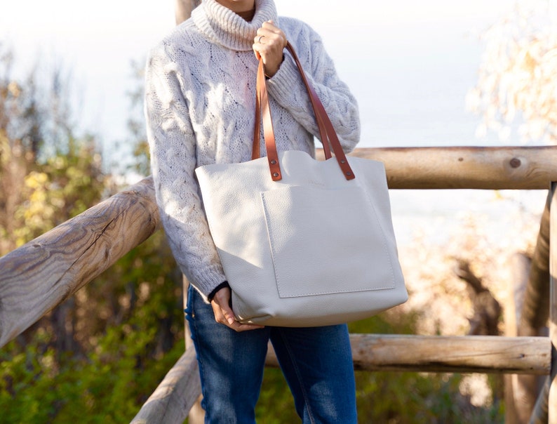 Watch the Tote Glo Bag. This tote bag is extremely light, the leather is ultra soft, buttery, very durable and resistant.
This bag is white, have tan leather handles and outside pocket. The bag appears grabbed by a model in front of wooden logs.