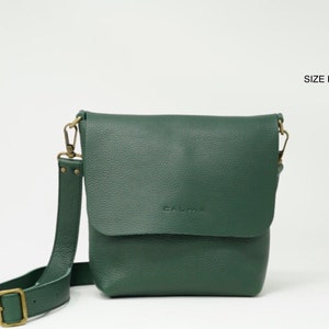 Watch the medium size UN, a leather crossbody bag with full grain leather, which feels very soft and silky.
This bag is forest green, comes on a white background and appears with its removable long strap hooked with old gold hardware.