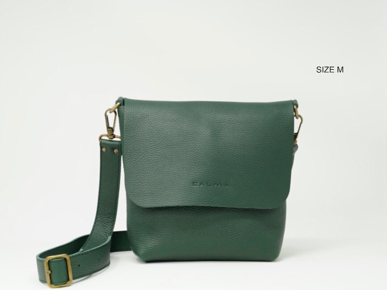 Watch the medium size UN, a leather crossbody bag with full grain leather, which feels very soft and silky.
This bag is forest green, comes on a white background and appears with its removable long strap hooked with old gold hardware.