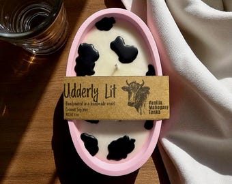 Może przedstawiać: Różowy owalny świecznik z białą świecą z czarnymi plamami krowy. Świeca ma brązową etykietę z napisem "Udderly Lit" i "Vanilla Mahogany Tonka". Świeca jest ręcznie wlewana do ręcznie robionego naczynia i wykonana z wosku sojowego kokosowego.