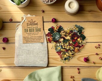 May include: A beige tea bath soak bag with a tag that reads "Velvet Expressions Forest Bloom Tea Bath Soak." A heart-shaped arrangement of dried flowers and herbs is next to the bag. The image is on a wooden surface.