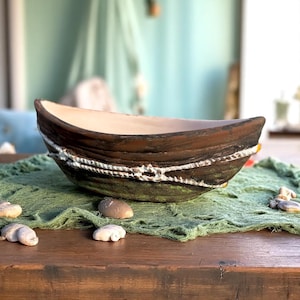 May include: A decorative boat-shaped ceramic bowl. The bowl features a brown exterior with a white rope detail and a green base. The interior is a light beige color. The bowl is displayed on a green mesh fabric.