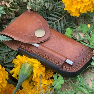 May include: A brown leather case with a metal button closure and a stitched design. The case is lying on a bed of green grass and yellow flowers.