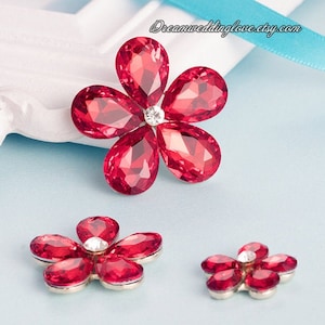 May include: Three red rhinestone flower embellishments of varying sizes. The largest flower has five petals and a clear rhinestone center. The two smaller flowers have five petals and a clear rhinestone center.