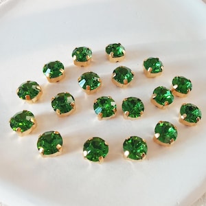 Op de afbeelding: Een verzameling van 16 groene strass steentjes met gouden settings. De strass steentjes zijn in een cirkel op een wit oppervlak gerangschikt.