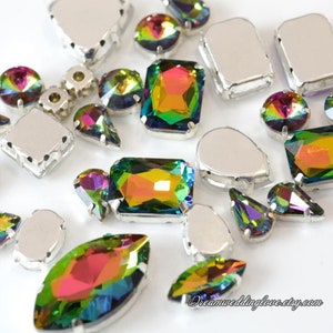 May include: Assortment of iridescent rainbow coloured rhinestones in various shapes and sizes, including round, oval, teardrop, and rectangular. The rhinestones have a silver backing.