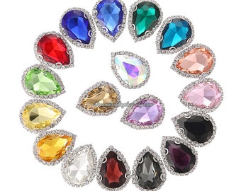 May include: A collection of teardrop-shaped rhinestones in various colors, including red, blue, green, and pink. Each gem is surrounded by a silver-colored, crystal-studded border. The gems are arranged in a circular pattern against a white background.