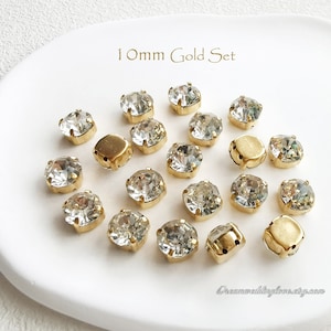 以下が含まれることがあります： 金色の枠で囲まれた透明なガラス製のラインストーン20個のセット。ラインストーンは直径10mmで、白い皿の上に円形に並べられています。皿には「10mm Gold Set」という文字が印刷されています。