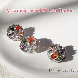 May include: Three silver-toned buttons with a microencrusted zircon design. Each button features a circular shape with a cluster of colourful stones in the centre. The stones are a mix of red, orange, yellow, green, purple, and clear. The buttons are 11.5 mm in diameter. Microencrusted Zircon Button.