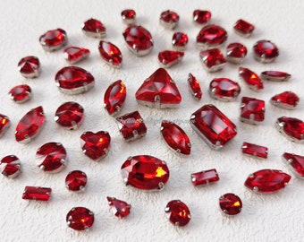 May include: Assortment of red glass rhinestones in various shapes and sizes, including round, oval, rectangular, and triangular. The rhinestones have a silver backing and are scattered on a white surface.